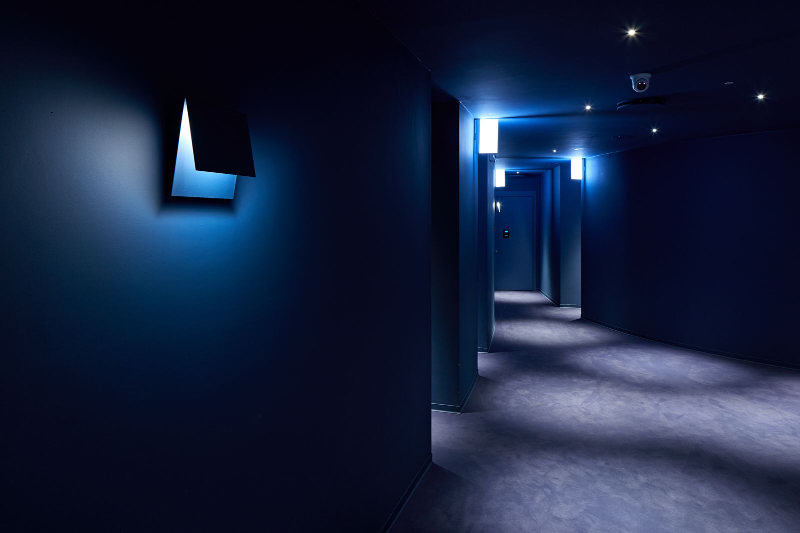 A dimly lit hallway with a blue hue, leading to a door at the end.