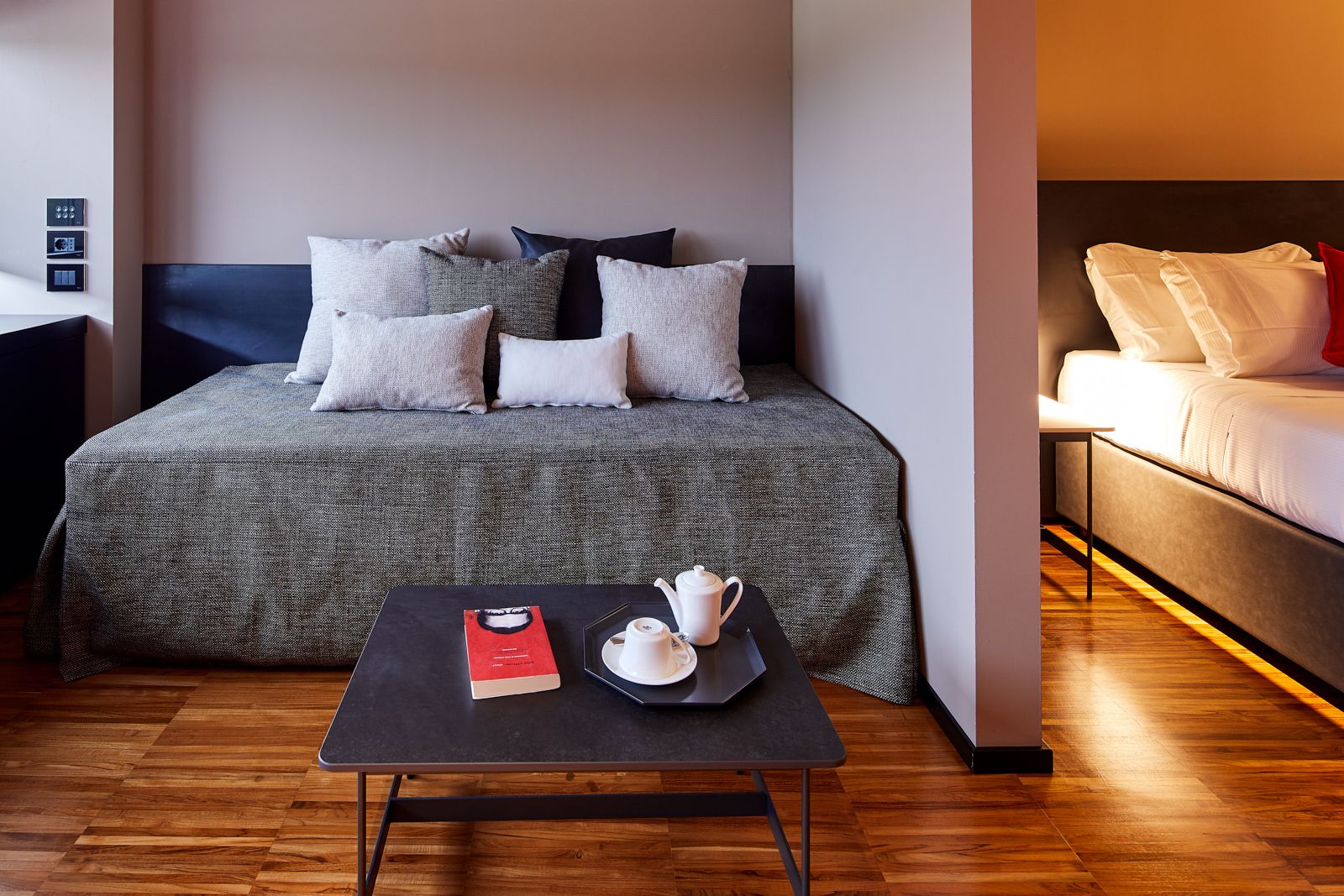 Una habitación de hotel moderna con una cama grande, otra más pequeña, una mesita con un libro y una tetera, y un termostato en la pared.