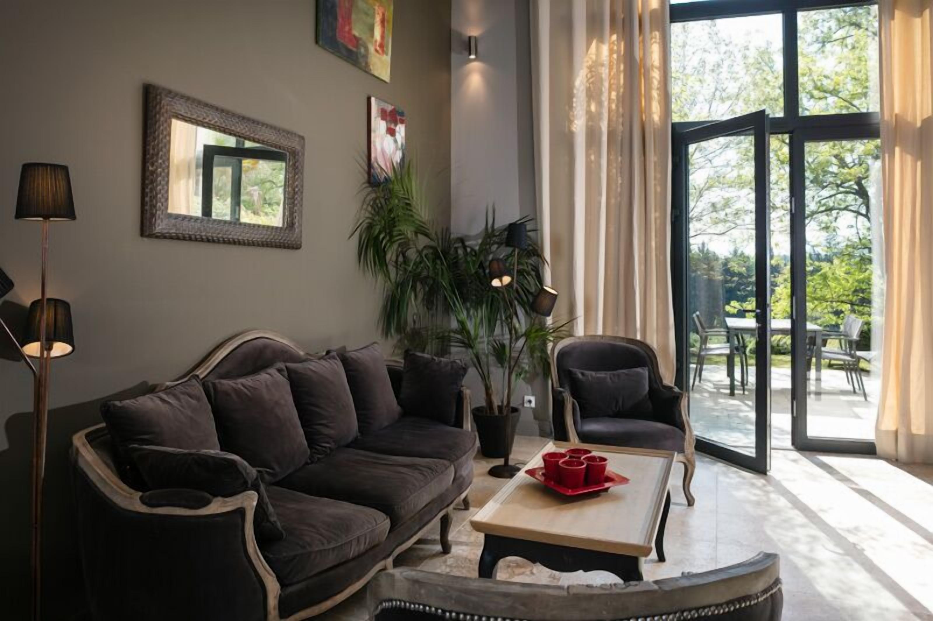 Un salon moderne avec un canapé marron foncé, une table basse sur laquelle reposent des tasses rouges, une plante en pot et une grande fenêtre donnant sur un paysage verdoyant.