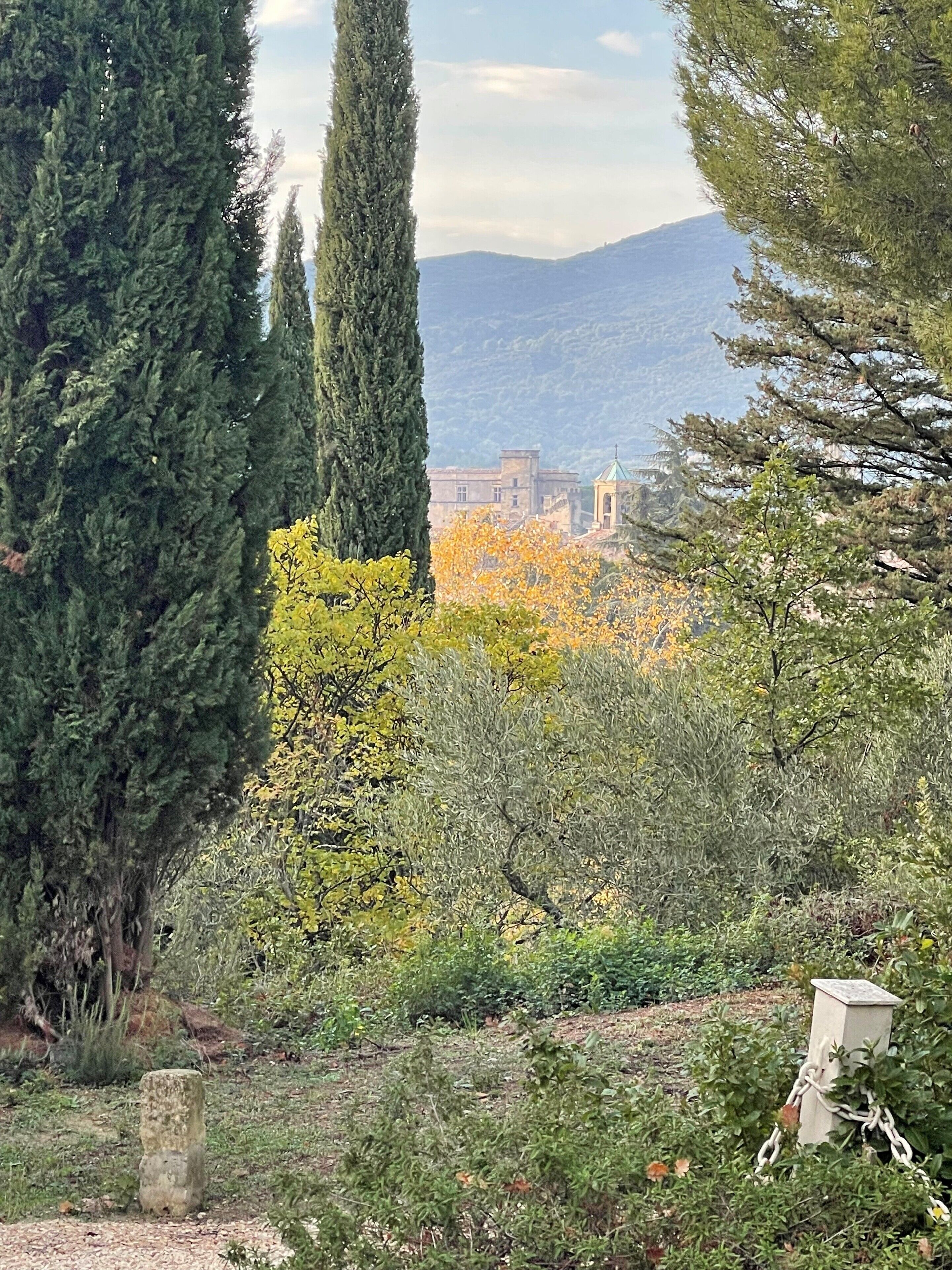 Un paisaje con altos cipreses, un pilar de piedra y un edificio con tejado verde al fondo.