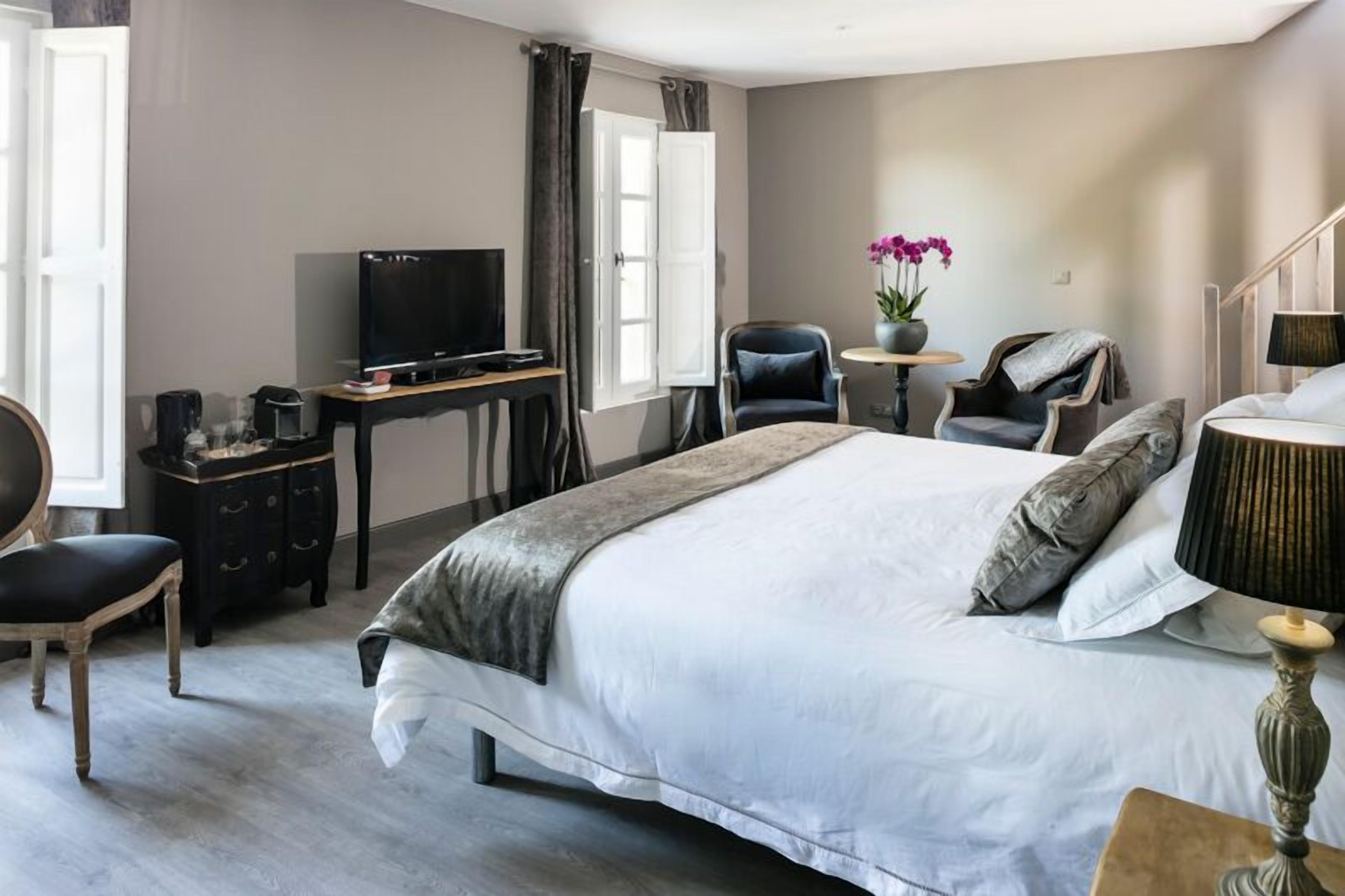 Une chambre avec un grand lit, un bureau avec une télévision, une chaise, une petite table avec une lampe et une fenêtre avec des rideaux.
