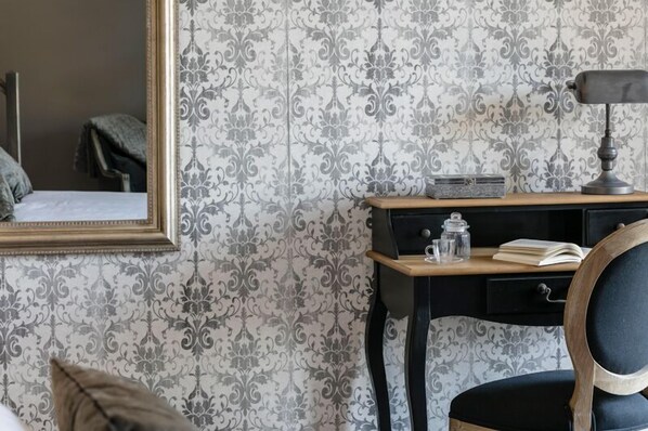 Una habitación con papel tapiz estampado, un escritorio de madera con lámpara, una silla y una cama.