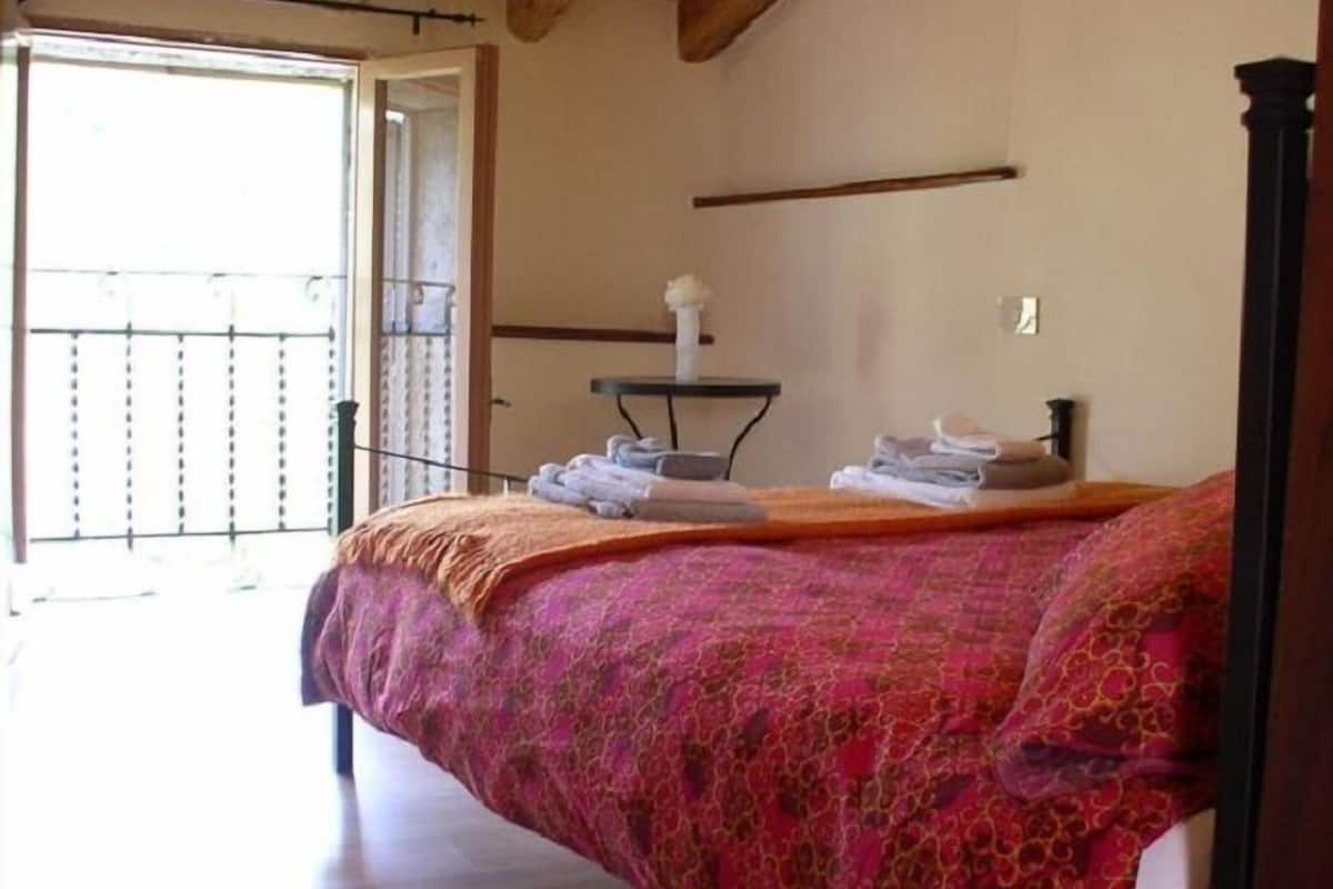 A bedroom with a wooden ceiling, a bed with a patterned blanket, a small table with towels, and a balcony door.