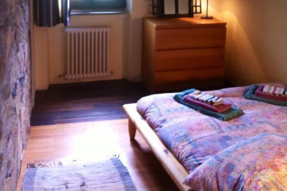 A bedroom with a wooden bed, a dresser, a window with curtains, a radiator, and a lamp.