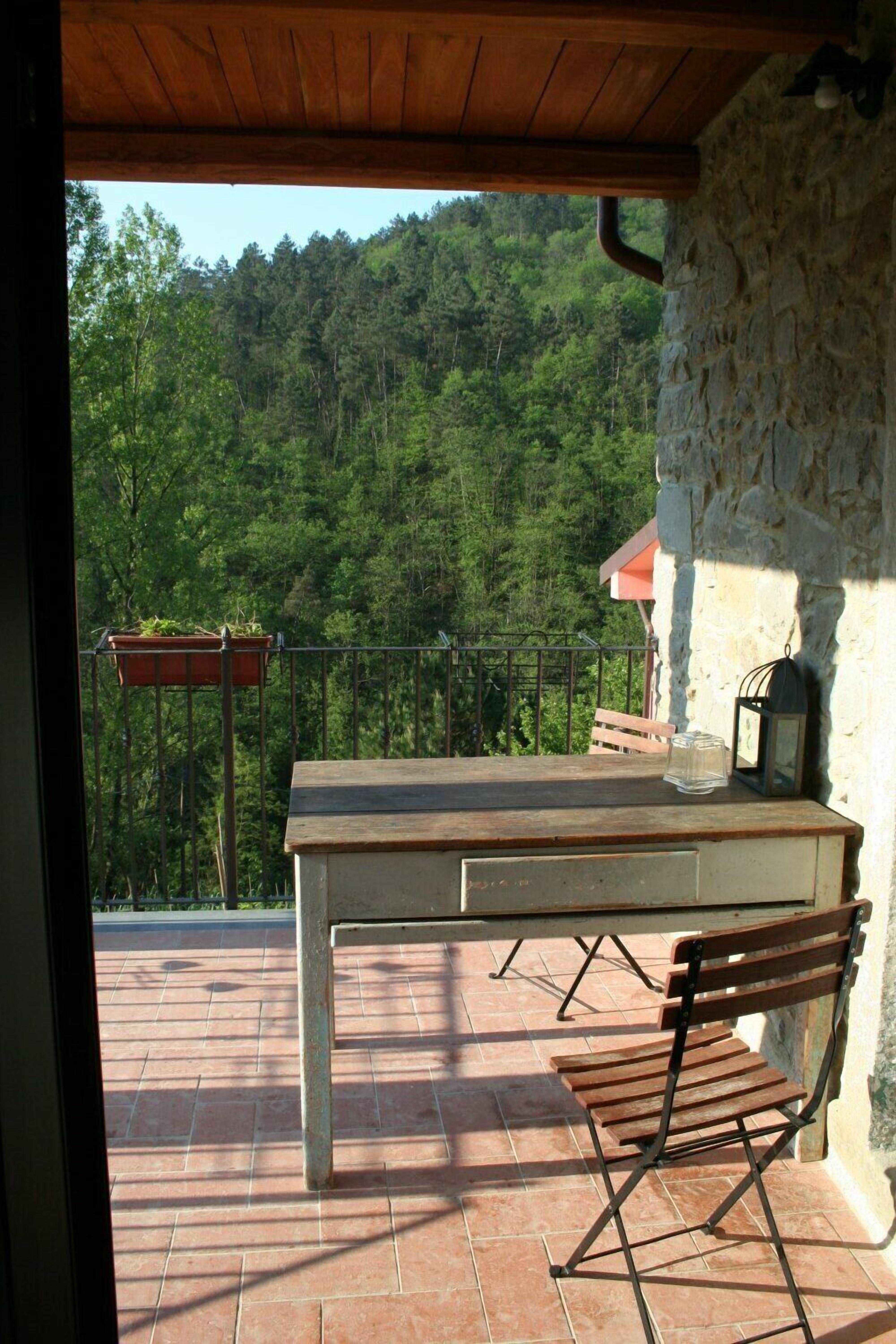 Mesa e banco de madeira num terraço com parede de pedra e vista para uma encosta arborizada.