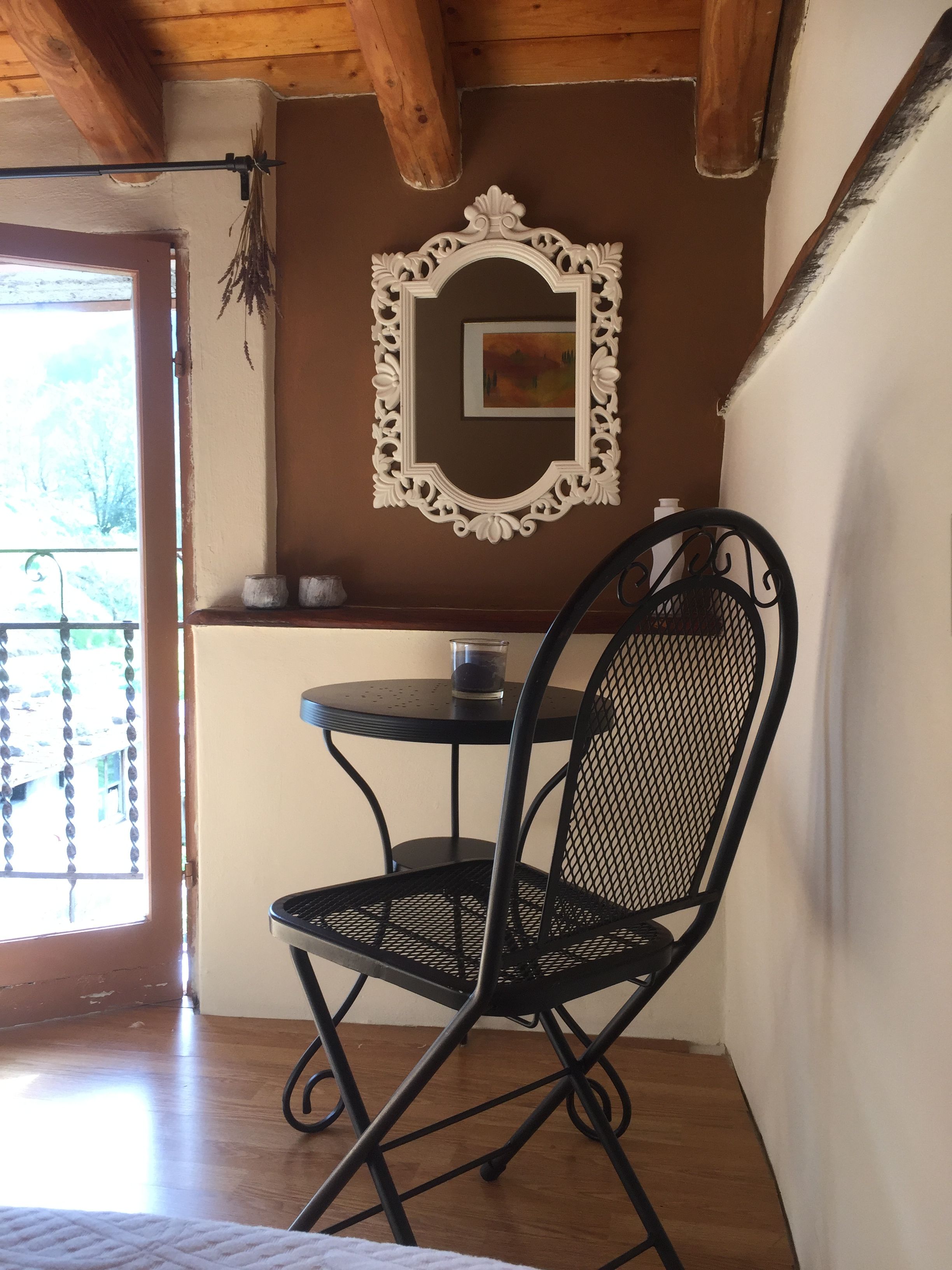 Um espaço de refeições pequeno, com uma cadeira e mesa de metal preto, um espelho decorativo e uma janela com vista para árvores.