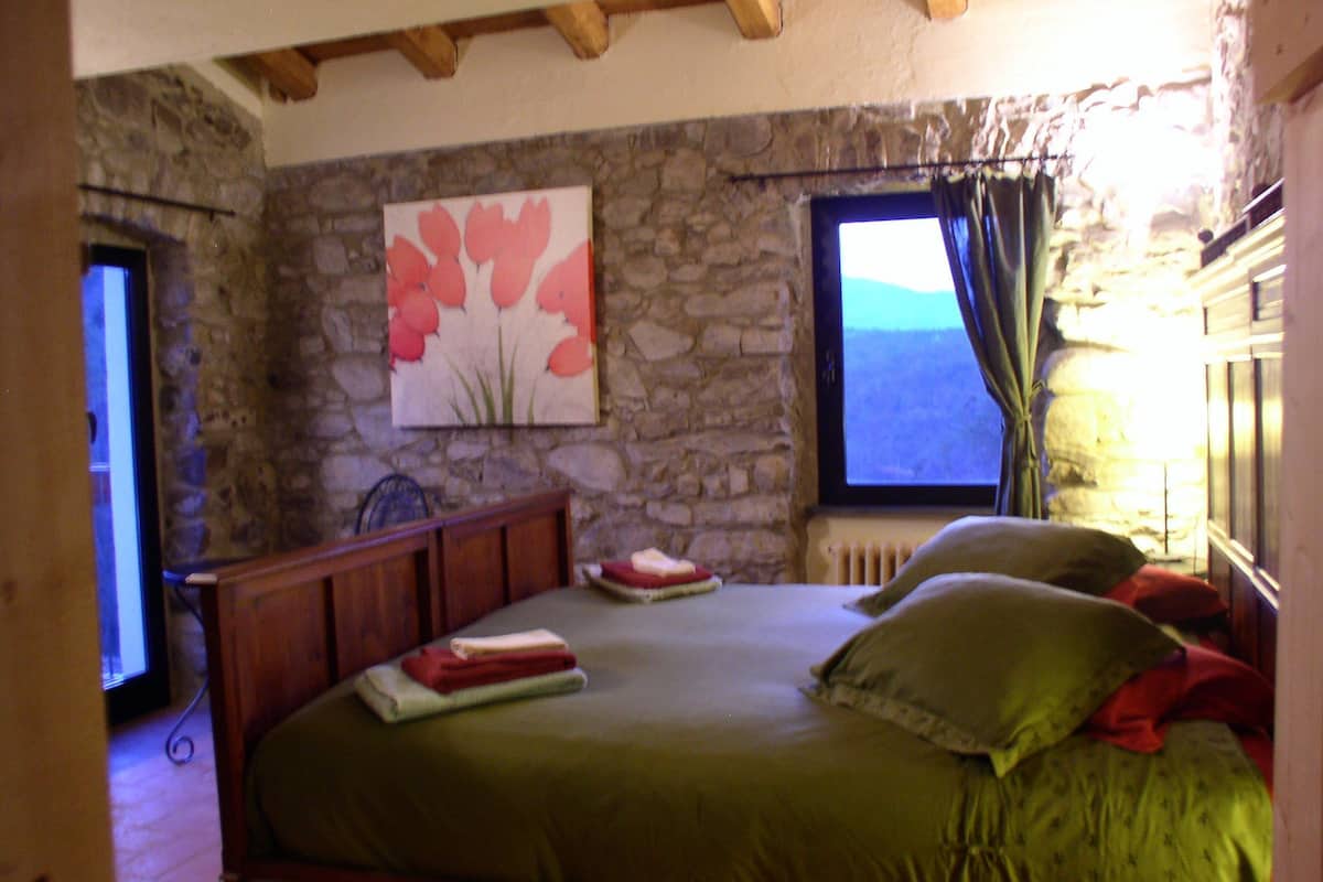 A bedroom with a stone wall, a bed with green bedding, a painting, and a window with a view.