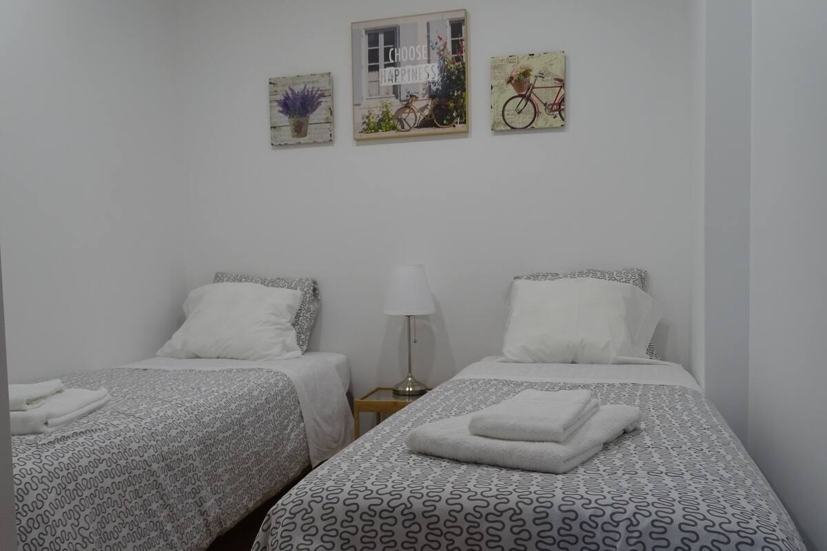 Une chambre avec deux lits, chacun agrémenté de draps et d’oreillers à motifs. Des œuvres d’art ornent les murs au-dessus des lits, notamment une représentation d’un vélo et une plante.