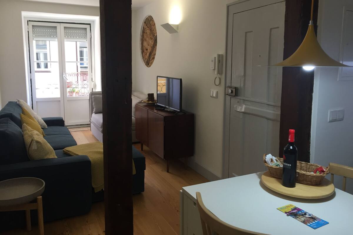 Un salon comprenant un canapé, un téléviseur, une table à manger avec une bouteille et un panier, ainsi qu’une porte dotée d’un interrupteur.