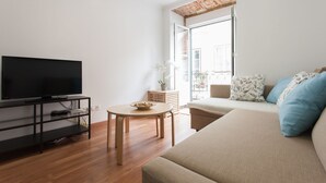 Un salon moderne avec un téléviseur à écran plat posé sur un meuble TV noir, une table ronde en bois sur laquelle se trouve un saladier, un canapé beige agrémenté de coussins à motifs et une grande fenêtre donnant sur l’extérieur.