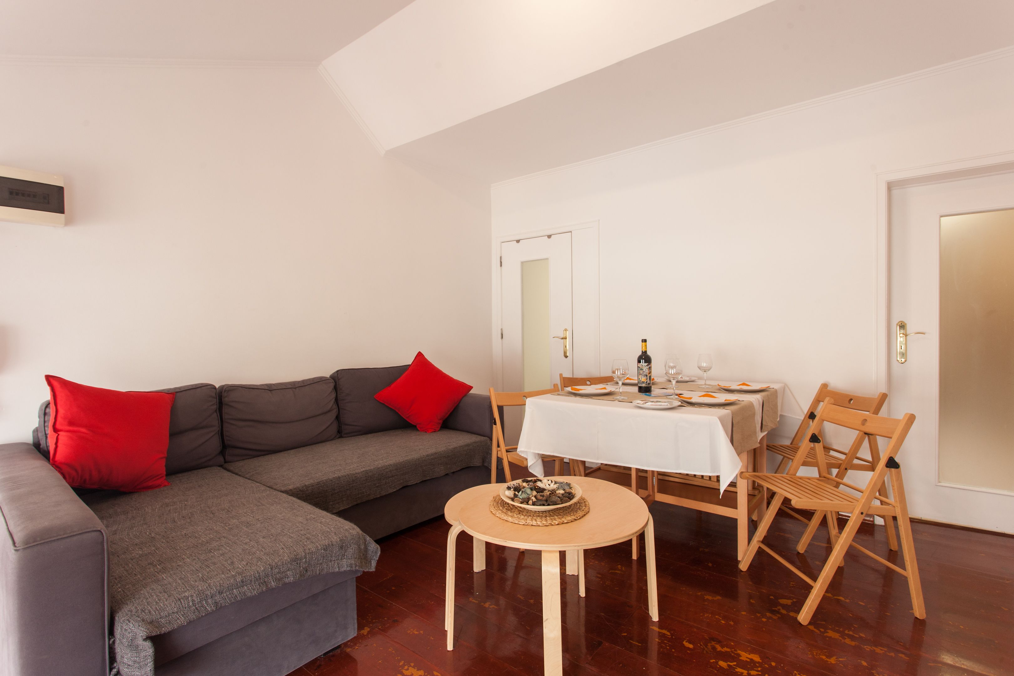 A living room with a grey sofa, red cushions, a round wooden table, and a dining table with chairs.