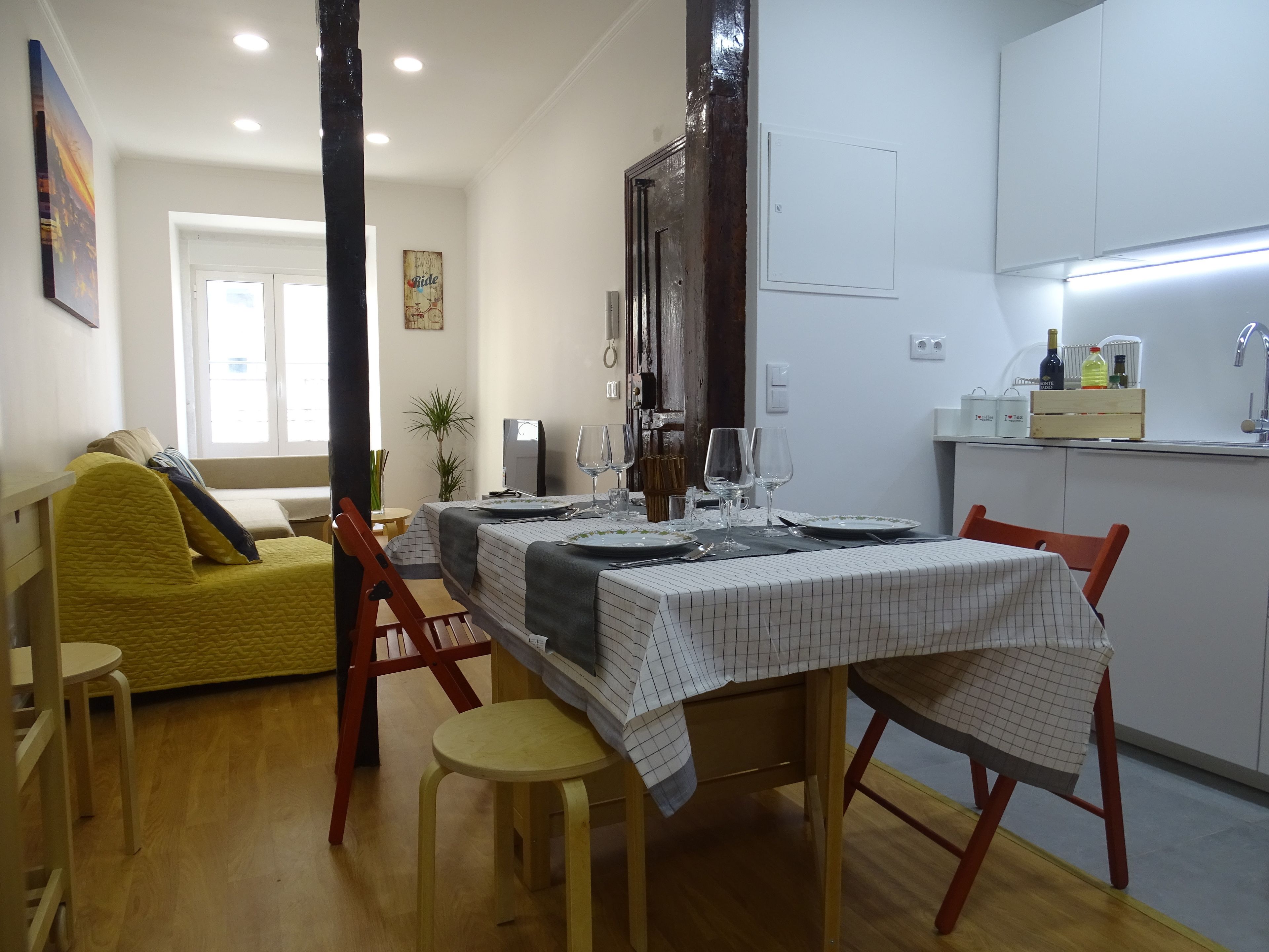 Un'area pranzo con un tavolo apparecchiato per quattro persone, circondato da sedie in legno. Si intravedono in secondo piano un divano e un piccolo tavolo con sedute.