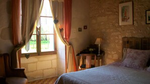 Un dormitorio con cama, una silla, una mesa con lámpara y un ventanal con cortinas.
