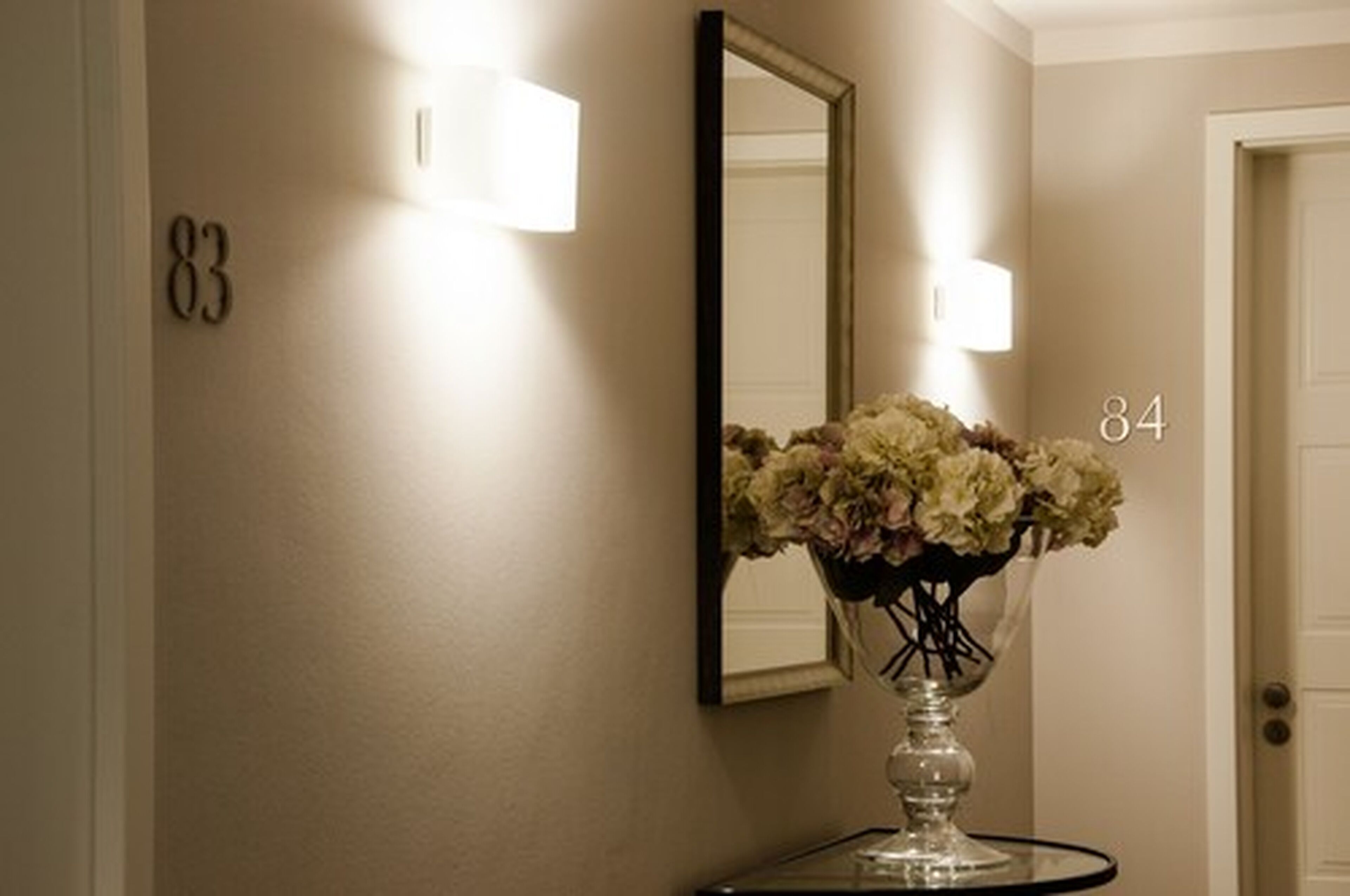 A hallway with a number 83 on the wall, a number 84 on the door, and a vase of flowers on a table.