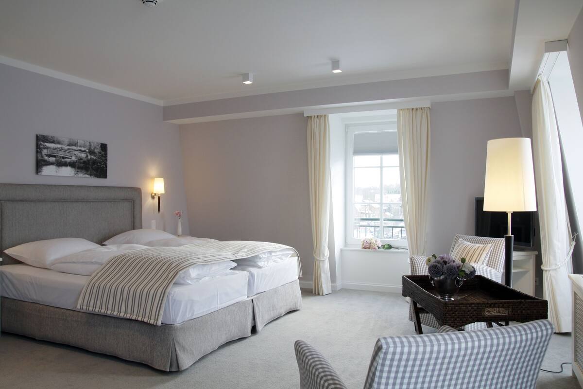 A hotel room with a large bed, a bedside table with a lamp, a chair, and a window with curtains.
