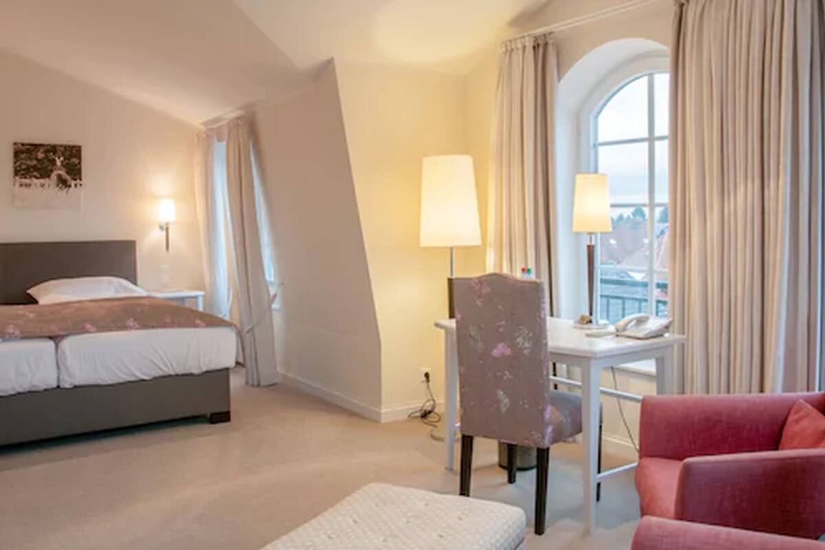 A hotel room with a bed, a desk with a chair, a red armchair, and a window with curtains.