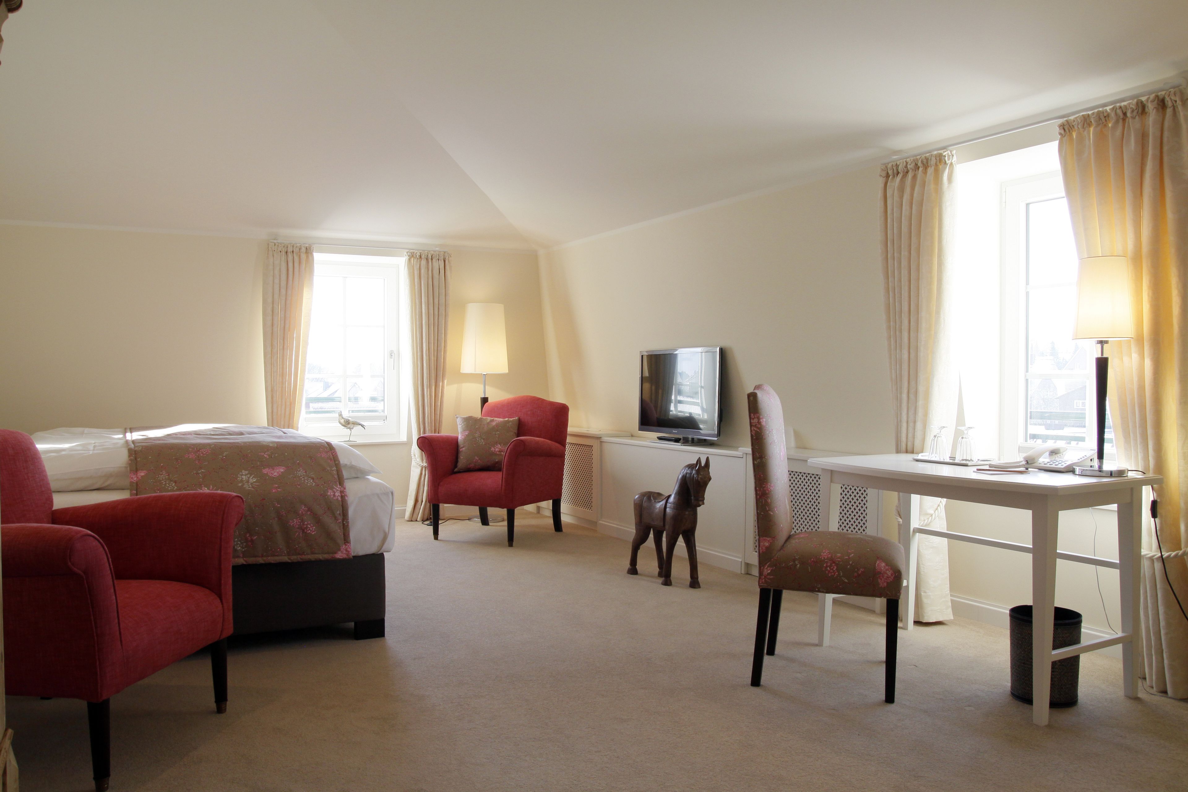Une chambre d’hôtel avec un lit, deux fauteuils rouges, un petit bureau avec une lampe, une télévision et une fenêtre avec des rideaux.