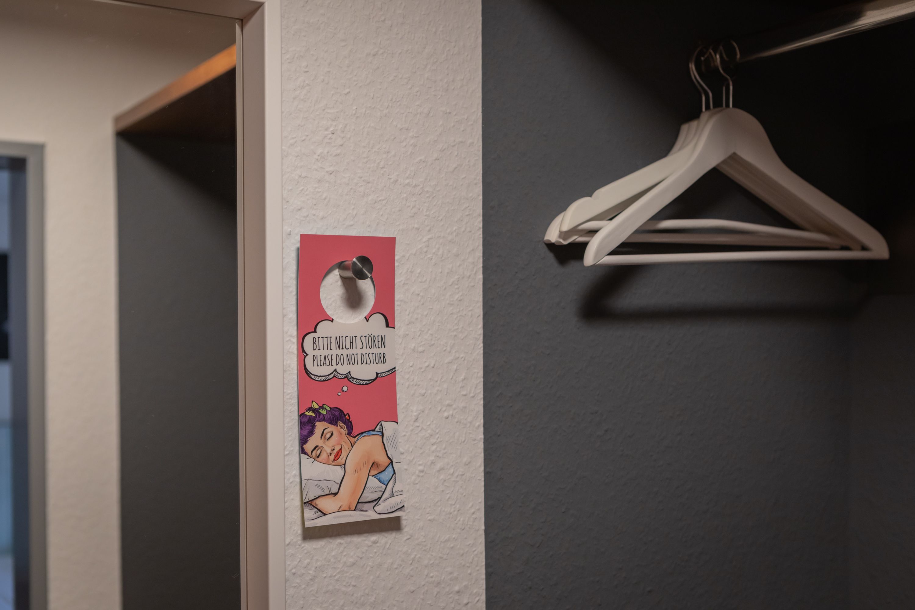 Ein an der Wand befestigtes Schild mit einer deutschen Beschriftung, ein weißer Kleiderbügel und eine teilweise sichtbare Tür.