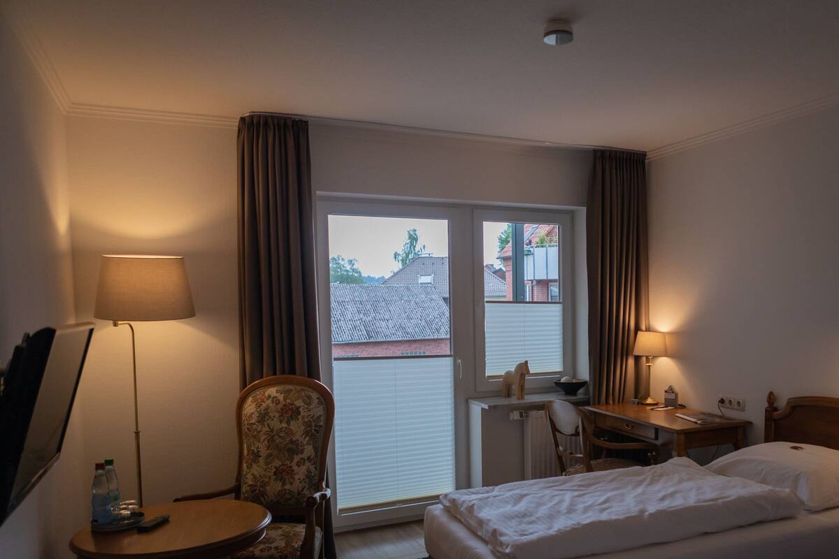 A hotel room with a bed, a desk, a chair, a lamp, and a window with curtains.