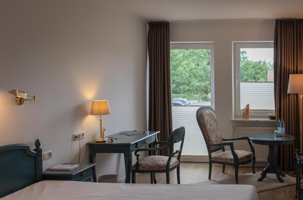 A hotel room with a bed, a desk with a lamp, two chairs, a small table with a lamp, and a view of the outdoors through a window.