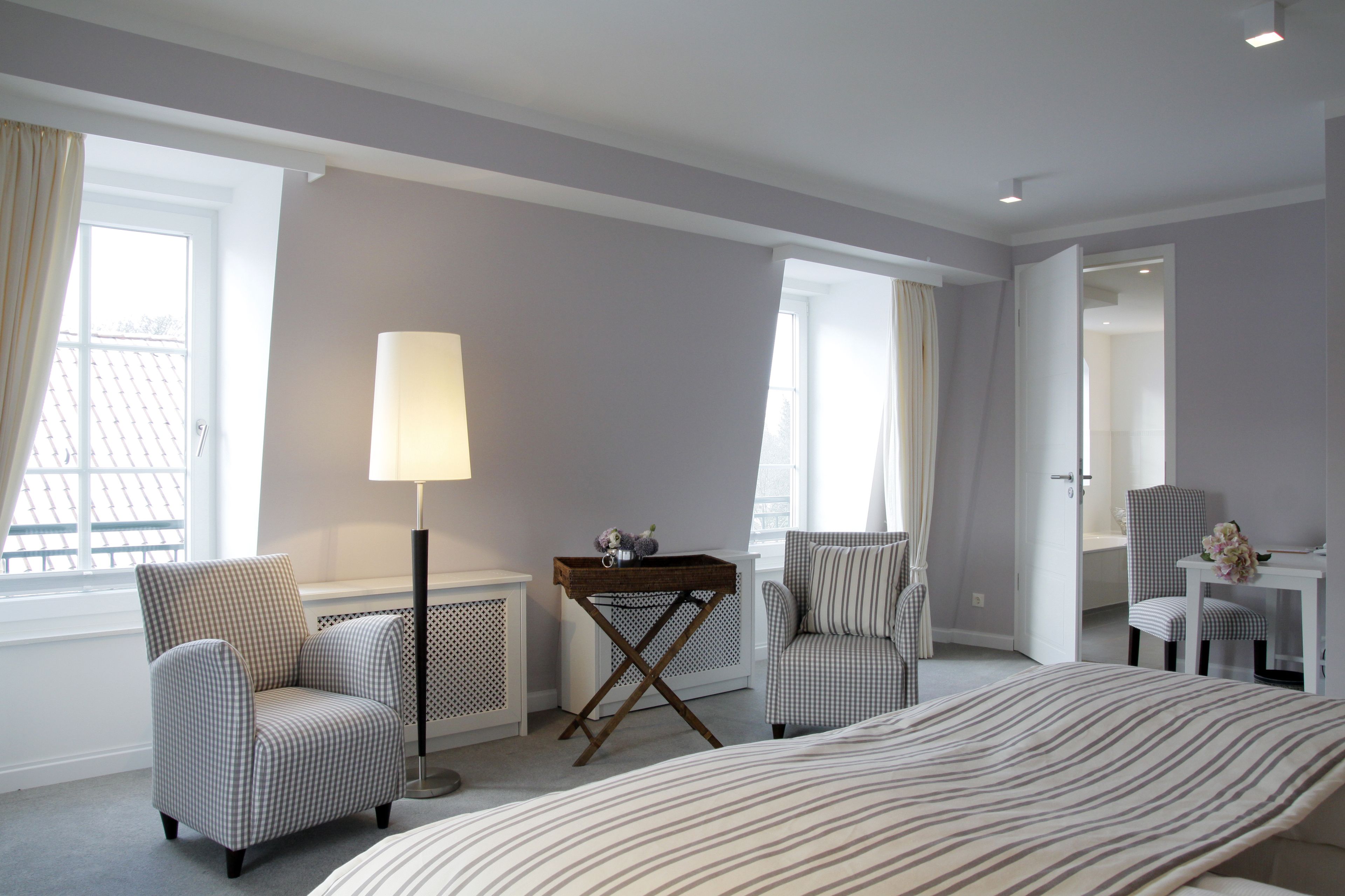 Habitación de hotel con cama, dos sillones, una mesita auxiliar, lámpara y vistas al exterior a través de amplias ventanas.