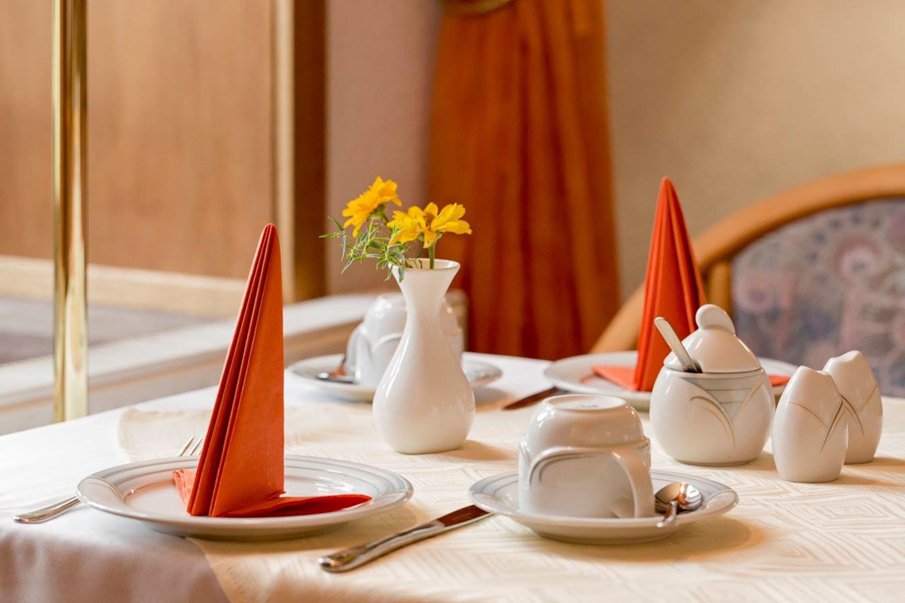 A neatly set dining table with a vase of yellow flowers, white teapots, and orange napkins.
