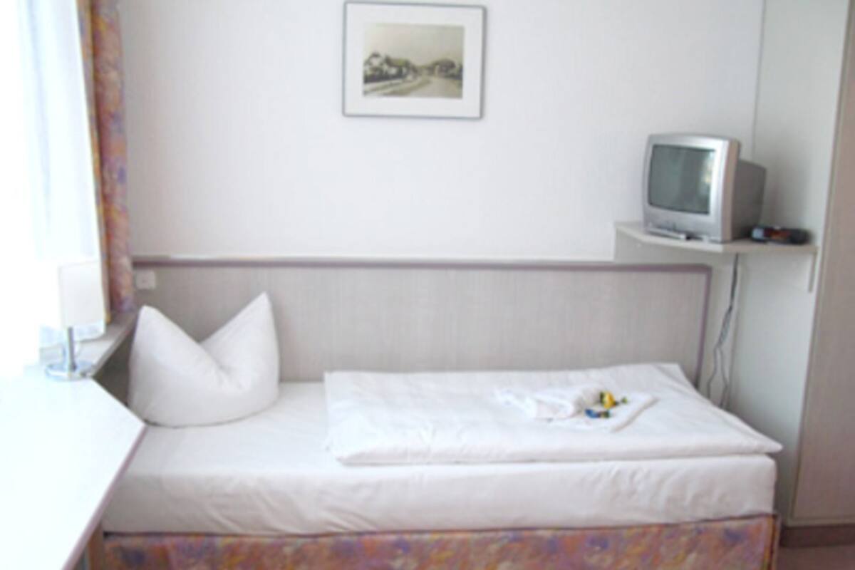 A small, neatly made bed with a white sheet and pillow, a television on a stand, and a framed picture on the wall.