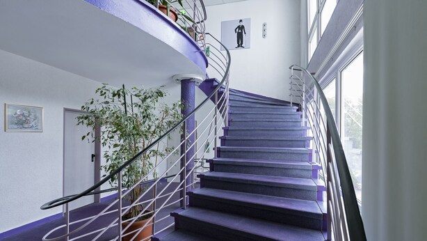 A spiral staircase with purple steps and metal railings, leading to a landing with a potted plant and a framed picture on the wall. - BIG Reisehotel Wolfen (Bitterfeld-Wolfen)