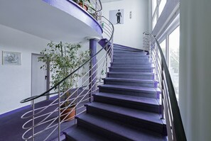 A spiral staircase with purple steps and metal railings, leading to a landing with a potted plant and a framed picture on the wall. - BIG Reisehotel Wolfen (Bitterfeld-Wolfen)