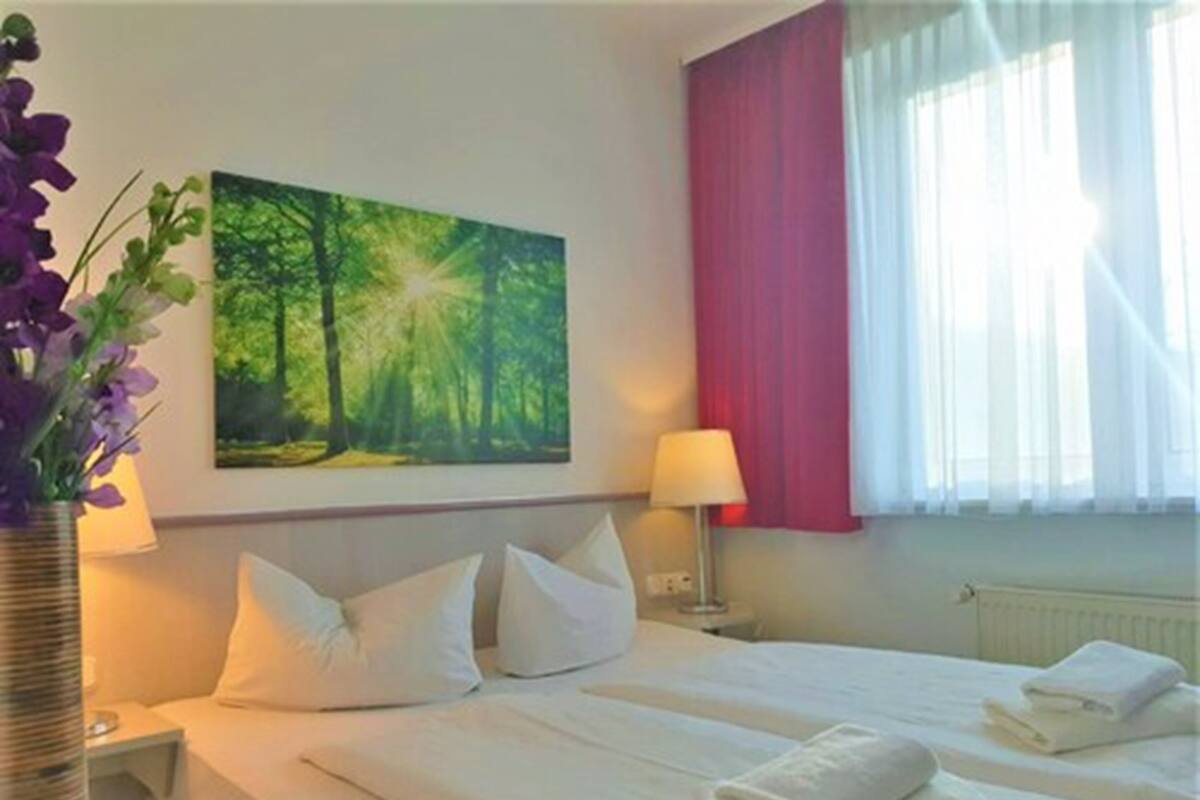 A hotel room with two beds, a painting of a forest, a vase with purple flowers, and a window with red curtains.