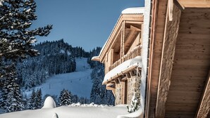 Un chalé de madera con base de piedra, rodeado de árboles cubiertos de nieve y una pista de esquí al fondo.