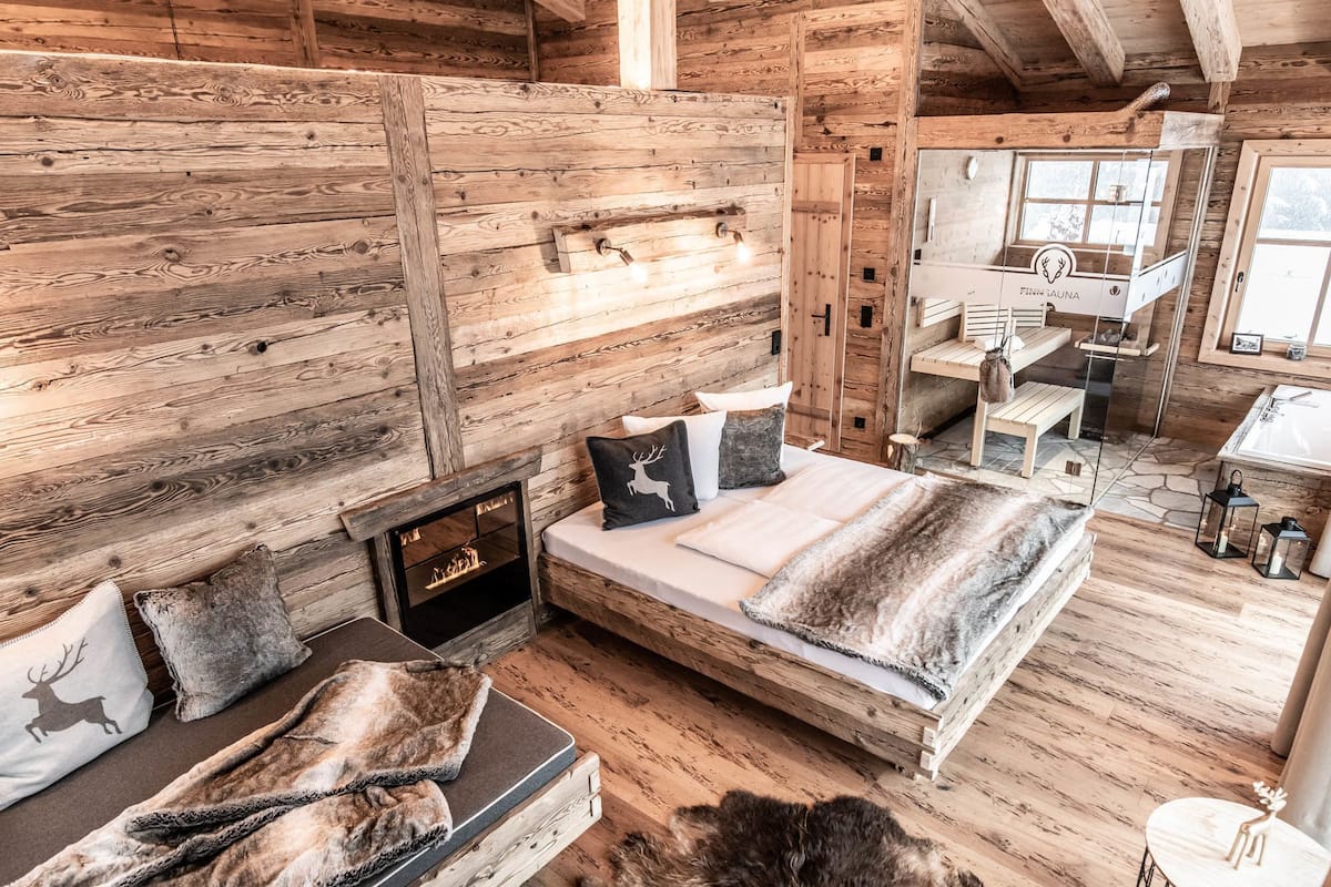 Interior rústico de una cabaña de madera con cama, chimenea y baño visible a través de un vidrio.