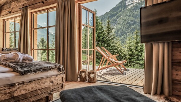 Una habitación de cabaña acogedora con una cama, una terraza de madera y un amplio ventanal con vistas a la montaña.