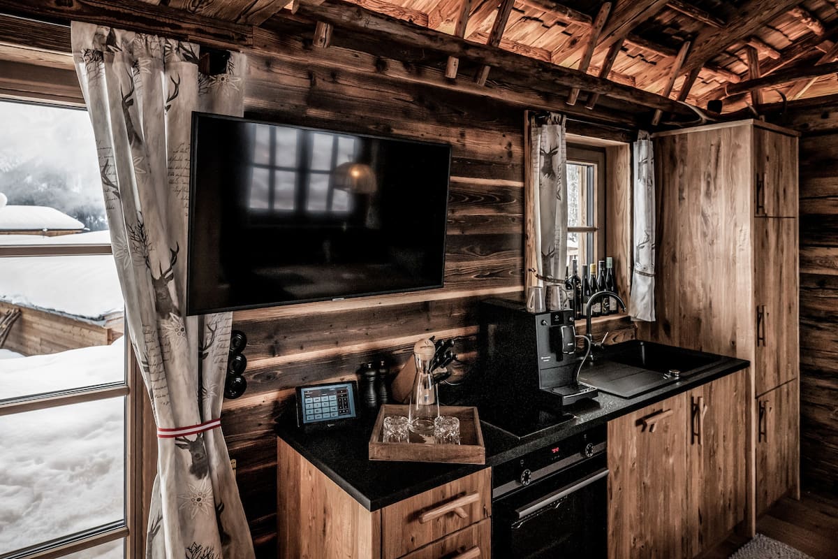 Interior rústico de una cabaña con detalles de madera, un televisor de pantalla plana grande montado en la pared, una zona de cocina con fregadero y encimera, y una ventana con cortinas.