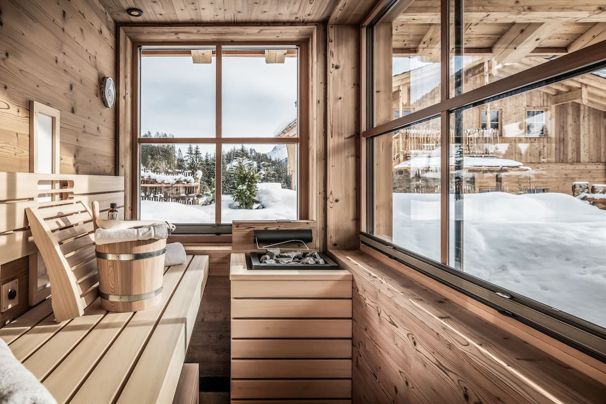 Interior de una cabaña de madera con sauna, bancos de madera y un ventanal con vistas a un paisaje nevado.