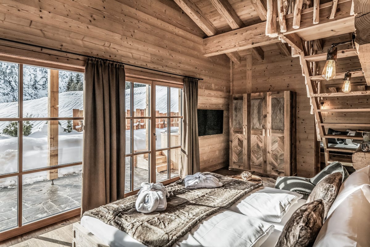Una habitación tipo cabaña de madera con un ventanal grande, vigas de madera, una cama con ropa de cama blanca y una chimenea.