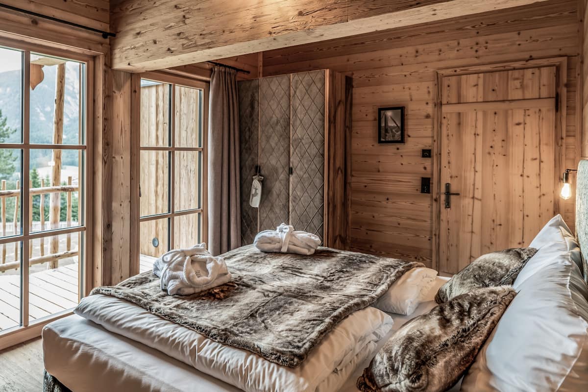 Un dormitorio de cabaña de madera con un ventanal grande, una cama con una manta de piel y un armario de madera.
