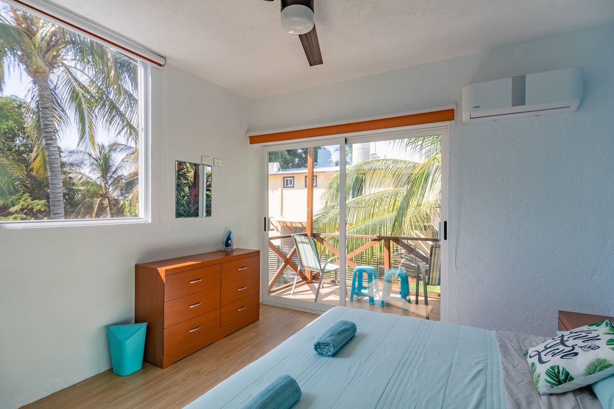 Un dormitorio con cama, cómoda, ventana y vista a un balcón con sillas y una palmera.