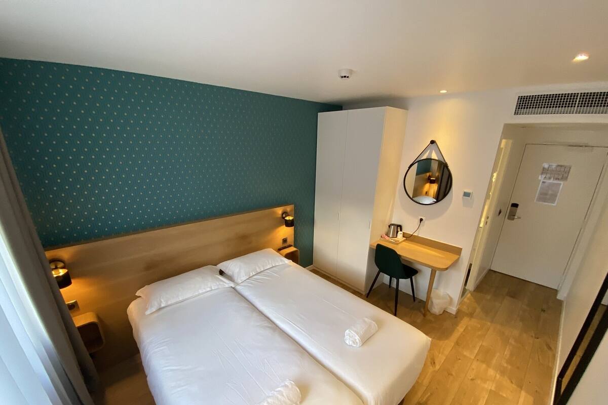 A hotel room with a double bed, a desk, a chair, a mirror, and a door.