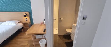 Une chambre d’hôtel comprenant un lit, une table en bois, une chaise, une petite poubelle et une salle de bain avec des toilettes et un lavabo.