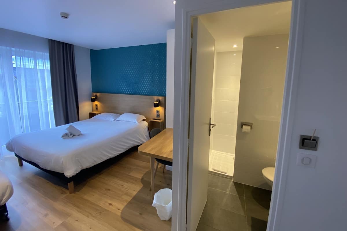 A modern hotel room with a large bed, bedside tables, a desk, and a bathroom visible through an open door.