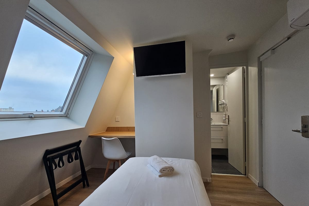A bedroom with a bed, a desk, a chair, a TV, and a bathroom visible through an open door.