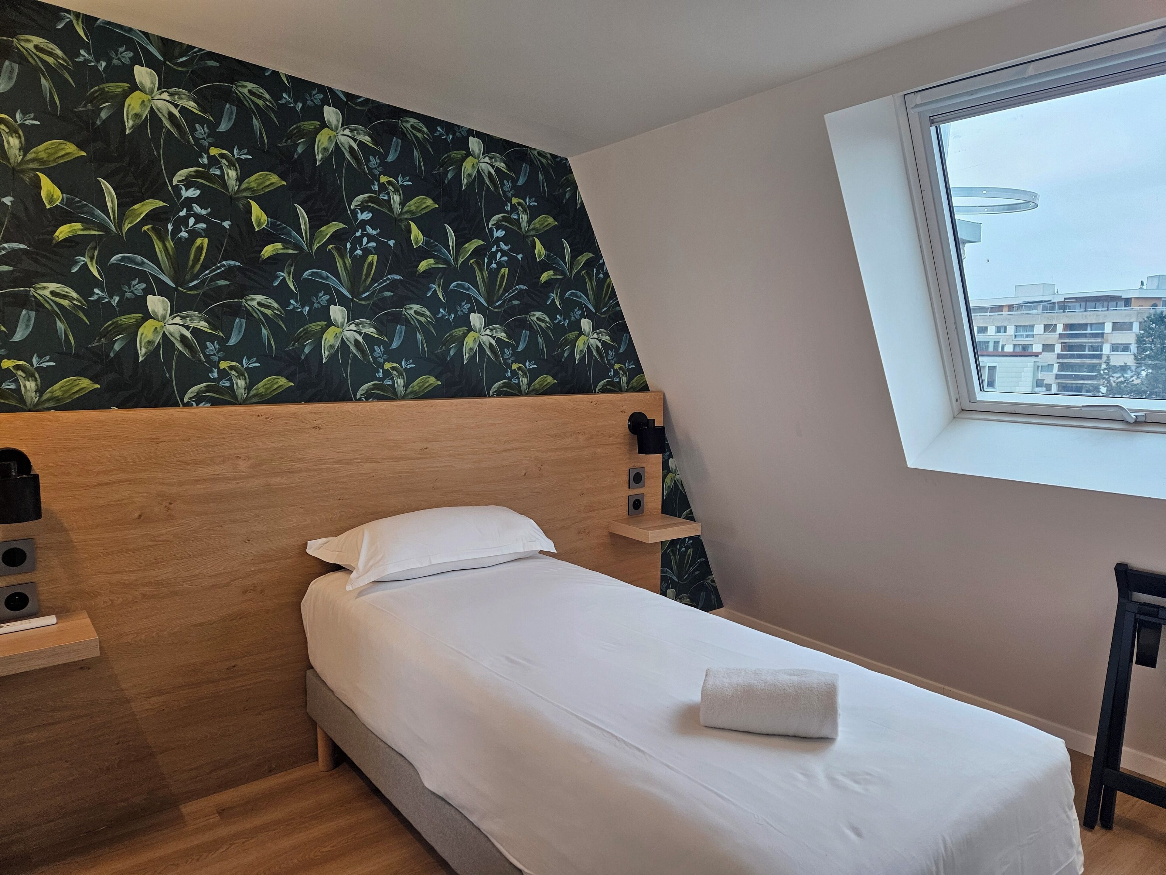 Una cama bien hecha con sábanas blancas y una toalla doblada, un cabecero de madera, una mesita de noche con una lámpara y una ventana con vista a edificios.
