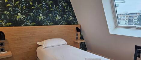 Una cama bien hecha con sábanas blancas y una toalla doblada, un cabecero de madera, una mesita de noche con una lámpara y una ventana con vista a edificios.