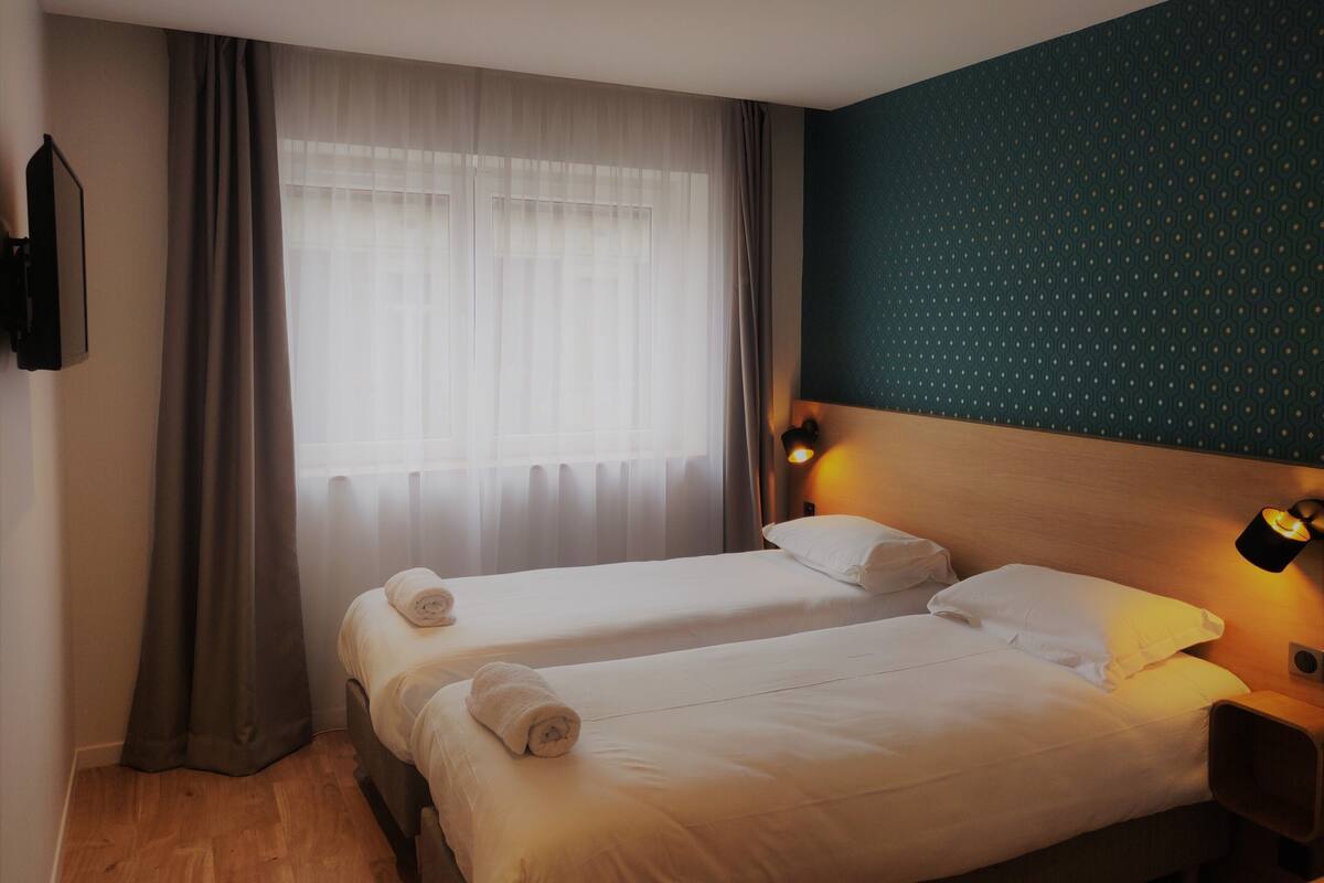 A hotel room with two beds, a large window with sheer curtains, a wall-mounted TV, and a patterned wall.