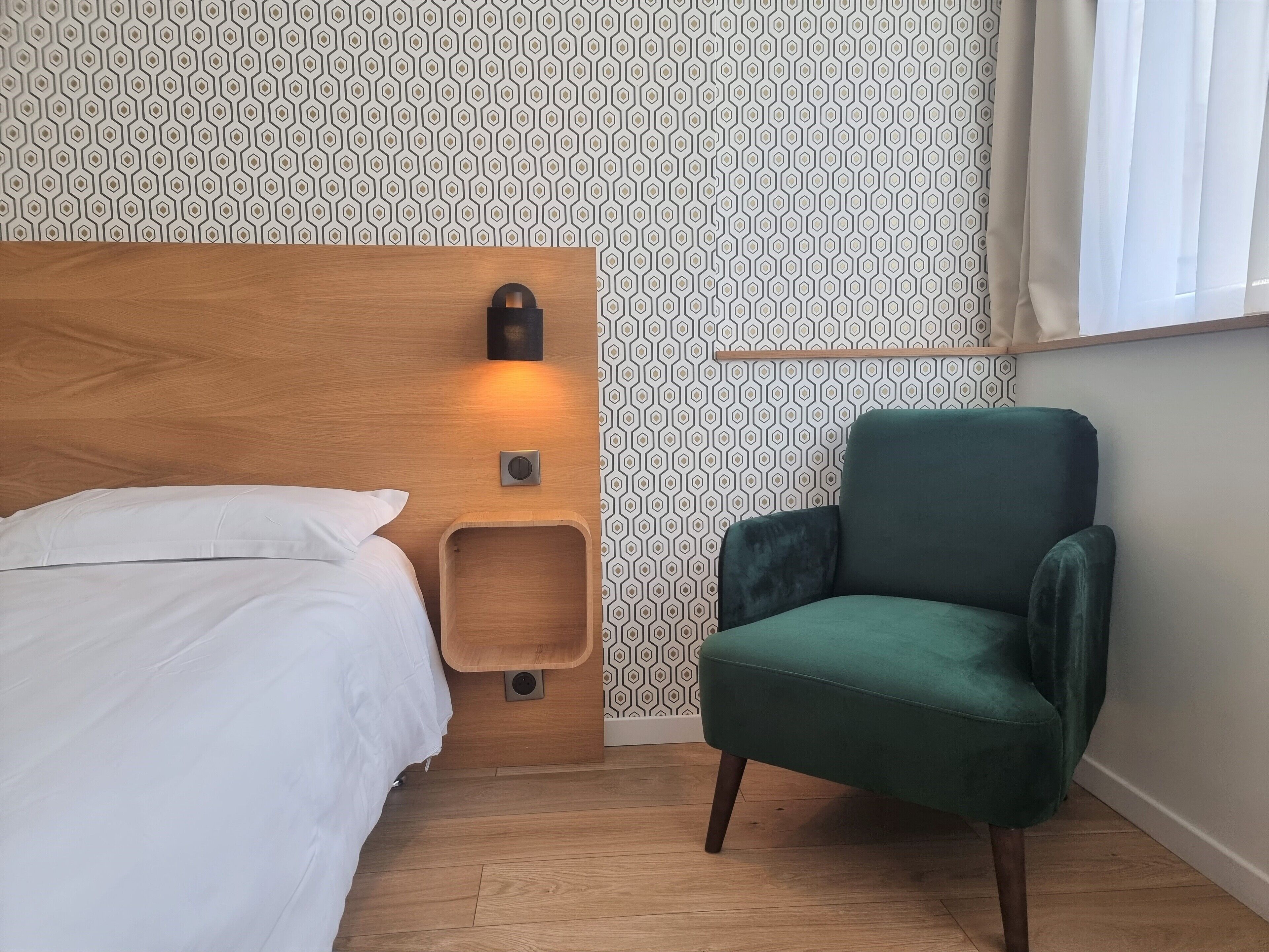 Habitación de hotel con cabecera de madera, un sillón verde, una mesita de noche y un aplique de pared.