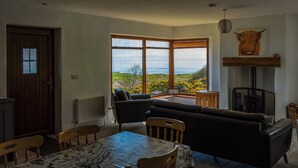 A living room with a view of the countryside, a fireplace, and a dining area with a table and chairs. - Makem's Self Catering Cottage (Ballycastle)