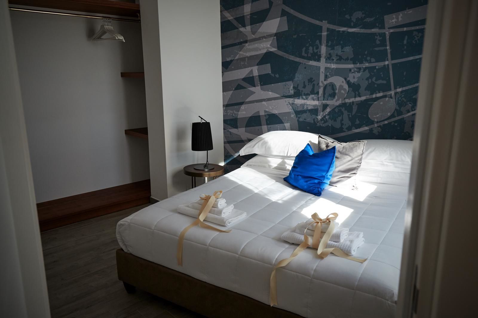 A neatly made bed with towels and a blue pillow, a small bedside table with a lamp, and a wall with a geometric pattern.