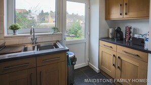 A kitchen with wooden cabinets, a sink, and a window overlooking a residential area. - Maidstone Family Homes - Fernhill (Maidstone)