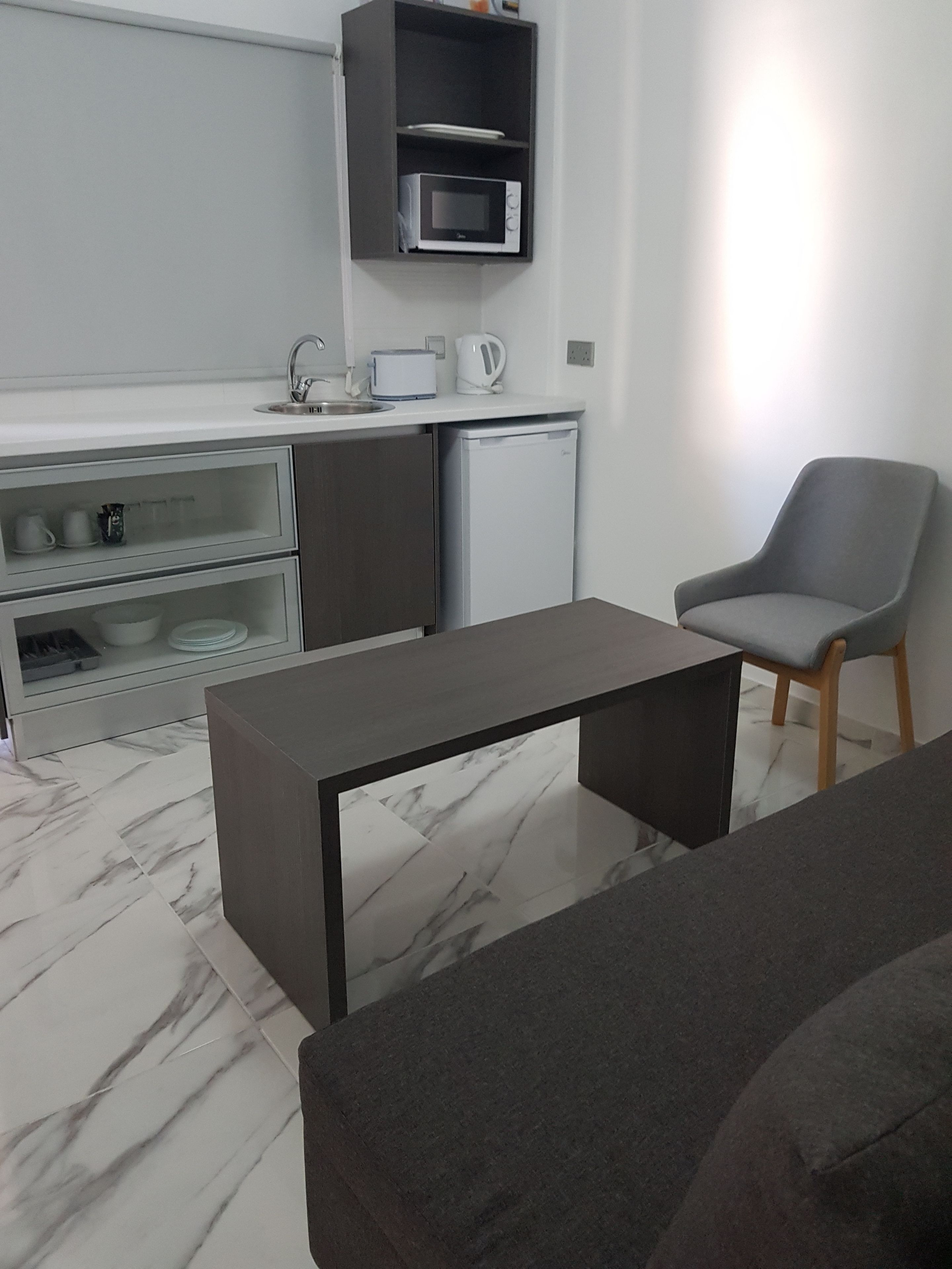 Una cocina moderna con microondas, hervidor y un refrigerador pequeño. En primer plano, hay una silla gris y una mesa oscura.